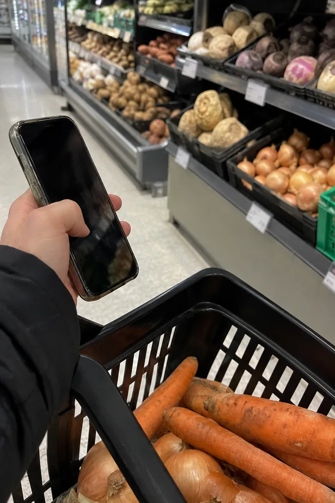 Käsi pitelee puhelinta ruokakaupan vihannesosastolla, korissa porkkanoita ja sipuleita